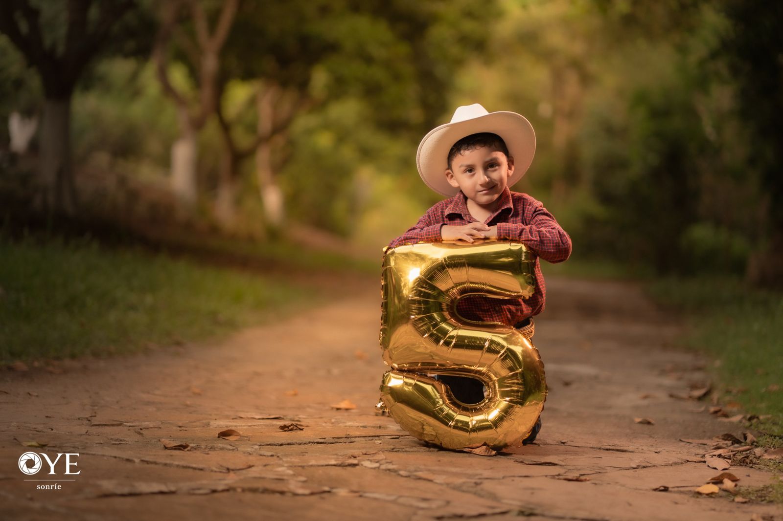 Niño con sombrero y globo dorado de número 5 en un camino entre árboles